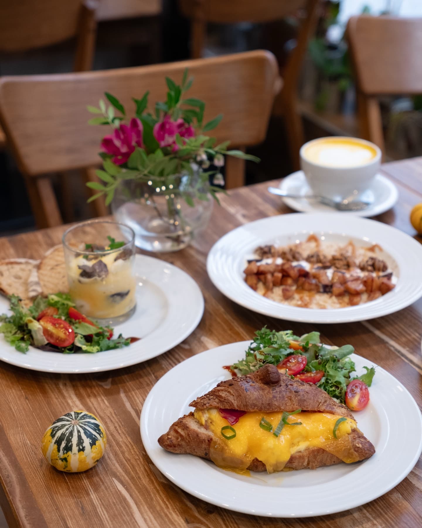 Brunch na dřevěném stole: croissant s vejci a cheddarem, salát, latte a kytička ve váze